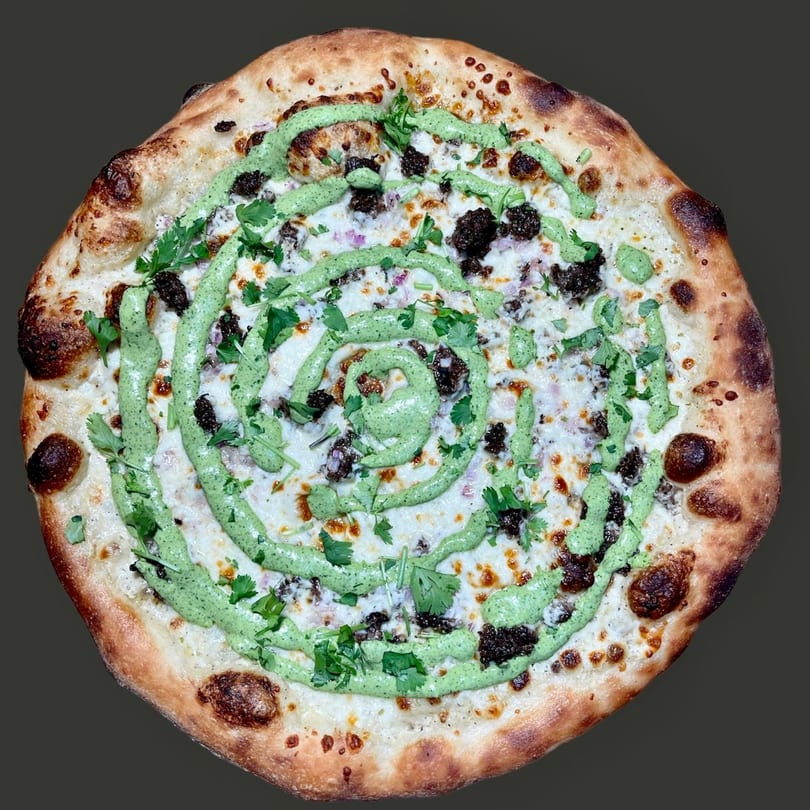 Round pizza with white sauce, dark crumbled toppings, red onion, green spiral sauce, and fresh cilantro.